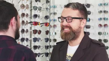 Olgun sakallı bir adam, parlak bir optik mağazasında bir dizi şık gözlükçüyle çevrili profesyonel bir optometristle canlı bir konuşma yaparken gülümsüyor. Atmosfer dost canlısı ve davetkar, uzman yardımını gösteriyor.