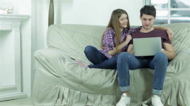 Neşeli genç bir çift, yumuşak, açık renkli bir kanepede sıcak bir anın tadını çıkarıyor, kahkahalar ve sıcaklığı paylaşıyorlar. Uzun saçlı ve neşeli bir gülüşü olan kız, dizüstü bilgisayarına odaklanmış erkek arkadaşının yanına eğiliyor ve bir yakınlık ortamı yaratıyor.