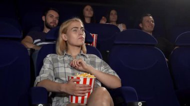 Sarı saçlı neşeli genç bir kadın, elinde patlamış mısırla gülümsüyor. Sıcak bir sinemada başparmağını kaldırıyor. Etrafı arkadaşlarla çevrili, atmosfer heyecan ve kahkahalarla dolu, bu da harika bir hafta sonu gezisi olmasını sağlıyor.