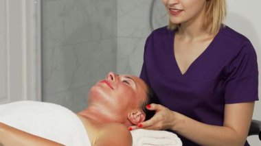 Yetenekli bir terapistin, gözleri kapalı mutlu bir şekilde gülümseyen rahat bayan müşterisinin kafasına nazikçe masaj yaptığı sakin bir sahne gözler önüne seriliyor. Yumuşak ışık ve sakinleştirici renklerle bezenmiş kaplıcanın sakin atmosferi...