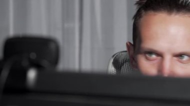 Yakın çekim, gece geç saatlerde bilgisayar ekranına dalmış bir erkeğin yoğun bakışlarını yakalar. Loş ışıklandırılmış çevre, modern bir profesyonelin adanmışlığını gösteren konsantrasyon ve kararlılık atmosferini artırır.