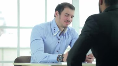 Biri açık mavi gömlekli, diğeri parlak siyah takım elbiseli iki kendine güvenen işadamı, parlak ve çağdaş bir ofisteki bir sözleşme üzerine odaklanmış bir tartışmaya giriştiler. Atmosfer profesyonellik ve işbirliği ile suçlanıyor.