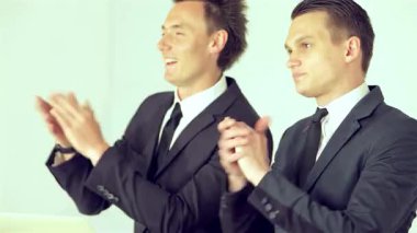 İki iyi giyimli iş adamı, biri şık saç stiline sahip, coşkulu bir iş konferansında bir konuşmacıyı coşkuyla alkışlıyor, pozitiflik ve bağlılık saçıyor. İfadeleri heyecan ve desteği yansıtıyor, işbirliği ruhunu somutlaştırıyor.