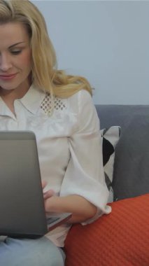 Canlı turuncu yastıklarla bezenmiş pelüş gri bir kanepeye kurulmuş laptopunda coşkuyla dalgalanan sarı saçlı neşeli genç bir kadın. Yumuşak yastıklarla ve modern dekorlarla dolu güneşli oda sıcaklık ve yaratıcılık yayıyor.