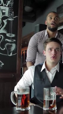 İki heyecanlı iş adamı, biri şık bir yelek ve diğeri kareli bir gömlek içinde canlı bir barda canlı bir şekilde TV 'ye tezahürat yapıyor ve bağırıyorlar. Atmosfer, buzlu bira bardaklarını kaldırırken enerjiyle doludur.