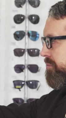 Neşeli sakallı bir adam, bir optometriste hevesli bir şekilde şık gözlükler seçerek merakını ve hevesini gösteriyor. Göz kamaştırıcı güneş gözlüğü ve klasik çerçevelerin yer aldığı göz kamaştırıcı gözlük dizisi,
