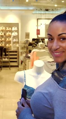 Zarif bir Afrikalı Amerikalı kadın modern bir butikte şık bir iç çamaşırı sergisinin fotoğrafını çekerken kendine güvenle ışıldıyor. Akıllı telefonuyla şık bir mankenin yanında duruyor. Moda zevkini ve alışverişin heyecanını somutlaştırıyor.