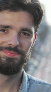 Şık sakallı şık bir genç adam, canlı bir şehir caddesine kurulmuş enerjik bir gülümseme sergiliyor. Rahat duruşu ve davetkar ifadesi, dinamik kentsel yaşam tarzına izleyicileri çeken güven ve neşe saçıyor.