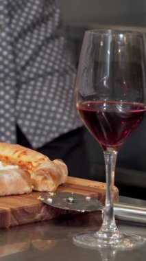 Genç bir adam heyecanlı bir şekilde ağız sulandırıcı bir pizzayı kapıyor, güzel bir şekilde ahşap bir tahtanın üzerinde düzenleniyor. Samimi daire, sahneyi hazırlayan bir bardak koyu kırmızı şarapla mükemmel bir şekilde eşleştirilmiş taze pişmiş pizza aromasıyla dolu.