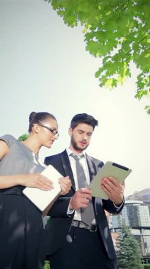 Kendine güvenen bir işadamı ve dengeli bir iş kadını, yeşillik dolu canlı bir şehir zeminine karşı hazırlanan parlak tabletlerinin mali özetlerini incelerken hareketli bir tartışma içine giriyorlar. Keskin profesyonel kıyafetleri ve odaklanmaları.