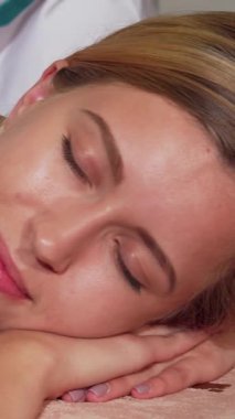 Huzurlu bir bayan müşteri, gözleri nazikçe kapalı ve yüzünde mutluluk dolu bir gülümseme, yumuşak ve rahatlatıcı renklerle kaplanmış lüks bir vücut masajına dalmış. Yetenekli masözlerin elleri ustalıkla derisinin üzerinde süzülerek huzurlu bir atmosfer yaratıyor.