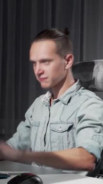 Sıradan bir kot gömlek giymiş, kendini işine adamış genç bir adam, gösterişli, modern bir masada oturur, gece geç saatlere kadar bilgisayar işleriyle uğraşır. Ortam ışığının yumuşak parıltısı sakin bir atmosfer yaratıyor, rahat duruşunu ve memnuniyetini vurguluyor.