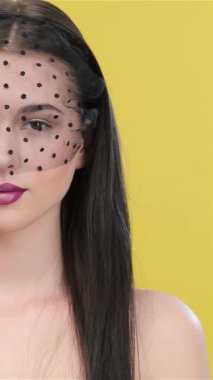 Büyüleyici Asyalı bir kadın, uzun, esmer saçlı, düşünceli bir şekilde avucunun içine bakıyor, narin özellikleri şık siyah bir peçeyle vurgulanıyor. Parlak sarı bir arkaplan karşısında, bu çarpıcı görüntü zarafet ve cazibe yayıyor, izleyicileri davet ediyor.