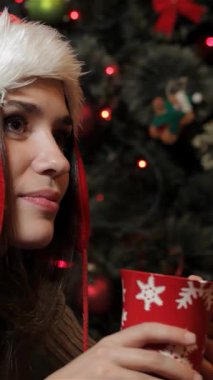 Neşeli, genç, beyaz bir kadın, Noel Baba şapkasıyla süslenmiş, dumanı tüten bir fincan sıcak kakaonun, sıcaklığın ve bayram neşesinin tadını çıkarıyor. Yanıp sönen ışıkların ve Noel ağacındaki canlı süslerin ortasında düşünceli bir şekilde bakıyor.