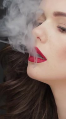 Bu büyüleyici ağır çekimde, dalgalı esmer saçlı çarpıcı beyaz bir kadın yoğun bir şekilde kameraya bakıyor, parlak cildi koyu siyah bir fonda parlıyor. Görüntü, zarif güzelliğiyle kıvrımlı dumanın tam zıttıdır.