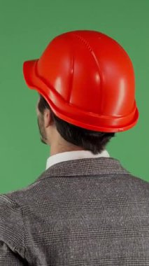 Gösterişli bir takım elbise ve parlak turuncu kasketli bir erkek mühendisin dikiz görüntüsü, canlı yeşil renkli bir ekrana bakarken hayal kırıklığı içinde boynunu ovuyor. Bu çarpıcı görüntü, yapıdaki hayal kırıklığının ve stresin ağırlığını özetliyor.