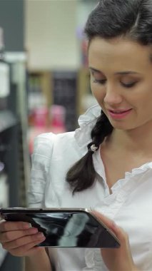 Neşeli genç bir kadın, ışıl ışıl gülümseyen bir gülümsemeyle canlı bir dükkanda şarap şişelerini karıştırıyor ve tabletini bilinçli seçimler yapmak için kullanıyor. Bu canlı sahne modern teknolojiyle mükemmel olanı seçmenin heyecanını harmanlıyor.