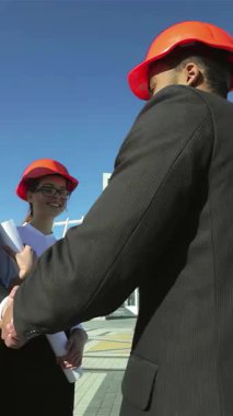 Gösterişli takımlar ve parlak turuncu şapkalar takmış, güneşli bir inşaat alanında coşkuyla el sıkışan, çığır açan fikirler alışverişinde bulunan kendinden emin mimarlardan oluşan bir üçlü. Bu dinamik sahne yaratıcılık ve işbirliği yayar.