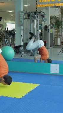 Orta Doğu kökenli odaklanmış genç bir adam, parlak, modern bir spor salonunda canlı sarı bir paspas üzerinde amuda kalkarak inanılmaz gücünü gösteriyor. Bu çarpıcı poz sadece atletik hünerini göstermiyor aynı zamanda kendini adamışlığını da gösteriyor.
