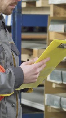 Koruyucu üniforması ve şapkası olan odaklanmış bir depo çalışanı elinde parlak sarı bir dosya tutarken envanterini titizlikle kontrol ediyor. Düzenli metal levhalarla çevrelenmiş bu sahne güvenlik ve hassasiyete olan bağlılığı somutlaştırıyor.