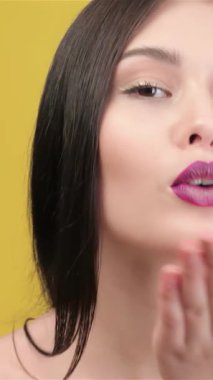 Parlak, koyu saçlı, hoş bir Asyalı kadın hava öpücüğü veriyor. Parlak gülüşü ve parlak mor dudakları, güneşli, sarı arka planda duruyor. Bu büyüleyici an onun neşeli ruhunu yakalıyor ve davetkar kişiliğini yaratıyor.