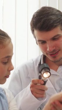 Parlak, hoş karşılayan bir pediatri ofisinde, koyu saçlı şefkatli bir erkek doktor özel bir cihaz kullanarak küçük bir kızın kolunu inceliyor. Saçı düzgün bir topuz şeklinde ve açık mavi bir elbise giyen kız, merakla izliyor.