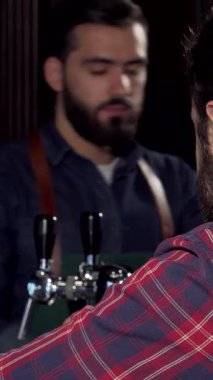 Keyifli bir erkek müşteri kameraya sırıtıyor, hareketli barda gıcır gıcır bir bira içiyor. Arka planda arkadaş canlısı bir barmen tarafından tamamlanan barın sıcak, davetkar parıltısı neşe ve dostluk dolu bir atmosfer yaratıyor.