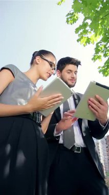 Zarif takım elbiseli kendine güvenen bir işadamı ve şık giyinmiş başarılı bir iş kadını, yemyeşil ağaçlar ve modern şehir mimarisi ile çevrili, açık mavi gökyüzünün altında tabletler kullanarak canlı bir tartışmaya girişiyor. Parlak gülümsemeleri ve...