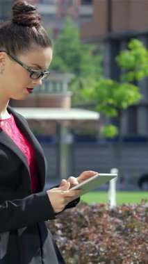 Şık siyah takım elbiseli, şık gözlükler takan dengeli genç bir iş kadını, açık hava ofis alanındaki hayat dolu meslektaşlarının arasında tabletine konsantre oluyor. Bu canlı sahne profesyonellik ve takım çalışması yayıyor, dinamiği vurguluyor.