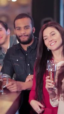 Canlı bir grup genç arkadaş, iki erkek ve iki kadın neşeli bir barda toplanıyor, bira bardaklarını kaldırırken coşkulu bir şekilde başparmaklarını kaldırıyorlar. Parlak gülüşleri ve bulaşıcı kahkahaları havayı heyecanla doldurup,