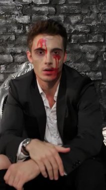 Büyüleyici bir erkek vampir, özel dikim siyah bir takım elbise giymiş, görkemli siyah bir kanepede uzanmış, delici bakışları ve kanlı yüzü yoğun bir Cadılar Bayramı havası yaratıyor. Bu çarpıcı görüntü, engebeli bir tuğla duvara karşı kurulmuş gotik bir görüntüyü temsil ediyor.