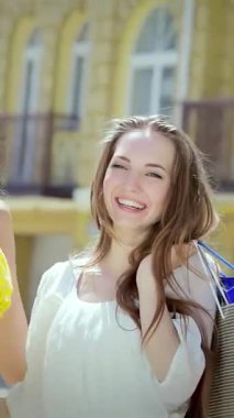 İki ışıl ışıl kız arkadaş, biri güneşli sarı elbiseli, diğeri rüzgarlı beyaz bluzlu, canlı şehir arkaplanına karşı canlı alışveriş torbalarını coşkuyla sergiliyorlar. Bulaşıcı gülümsemeleri ve kaygısız enerjileri arkadaşlığın heyecanını ve