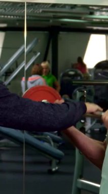 Dinamik bir spor salonu ortamında, kararlı gri saçlı yaşlı bir adam canlı kırmızı bir halteri kaldırır ve dikkatle pazularına odaklanır. Genç Kafkasyalı antrenörü yanında duruyor, cesaret verici tavsiyeler veriyor, modern bir atmosferi teşvik ediyor.