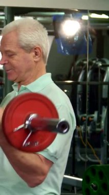 Dinamik bir spor salonu ortamında, kararlı gri saçlı yaşlı bir adam canlı kırmızı bir halteri kaldırır ve dikkatle pazularına odaklanır. Genç Kafkasyalı antrenörü yakınlarda duruyor, uzman tavsiyeleri ve teşvik sunuyor, motive edici bir atmosferi teşvik ediyor.