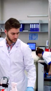 El değmemiş beyaz laboratuvar önlüğü giyen genç bir beyaz erkek bilim adamı laboratuvar ekipmanlarını titizlikle analiz ediyor. Kaşları yoğun bir şekilde çatılmış. Canlı laboratuvar çevresi ileri teknoloji ile uğulduyor, renkli ekipmanlar sergiliyor ve kendini adamış