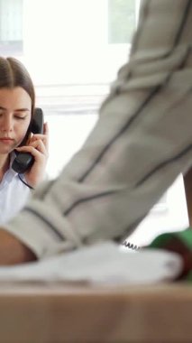 Renkli kumaşlar ve temel tasarım araçlarından oluşan bir kaleydoskop ile çevrili, canlı bir telefon konuşmasında odaklanmış genç bir bayan moda tasarımcısı ışınlar. Ön planda, kendini işine adamış bir meslektaş kumaşı bantla ustalıkla ölçer.