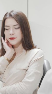 Endişeli bir ifadeyle genç bir kadın yanağına hafifçe dokunuyor, gösterişli, modern bir diş kliniğinde otururken diş ağrısını işaret ediyor. Bu sahne, parlak ve steril ortamda dişçi rahatsızlığının gerginliğini net bir şekilde yakalıyor.