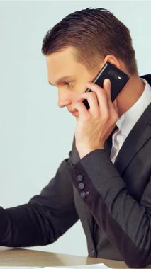 Özel dikim takım elbiseli kararlı genç bir işadamı, usta bir şekilde dizüstü bilgisayarında yazarak profesyonellik ve hırs yayarak bir telefon görüşmesi yapıyor. Parlak, modern ofis arkaplanı üretkenlik atmosferini yükseltiyor