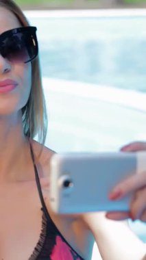 Canlı bikinili muhteşem genç bir kadın, parlak havuzun yanında poz verirken uzun saçları zarifçe akan neşeli bir selfie çeker. Neşeli bir gülümseme ve büyük güneş gözlükleriyle özgüven yayar ve kaygısız ruhunu somutlaştırır.