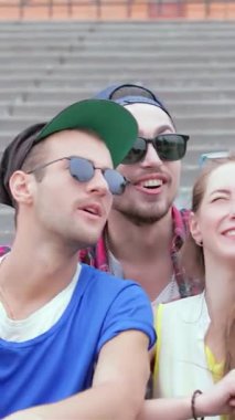 Genç arkadaşlardan oluşan canlı bir üçlü, bir erkek ve bir kadın, güneşli açık hava basamaklarında coşkulu bir şekilde selfie çekiyorlar, neşe ve dostluk saçıyorlar. Renkli kıyafetleri ve ışıl ışıl gülümsemeleri ölümsüzleştirmek için mükemmel bir atmosfer yaratıyor.