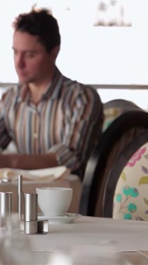 Çiçek aksanıyla süslenmiş şık bir kafede, genç bir işadamı tabletiyle etkileşime girerek lezzetli bir yemeğin tadını çıkarıyor. Sıcak ortam, zarif masa ortamları ve arka planda bir lokanta ile tamamlanmış.