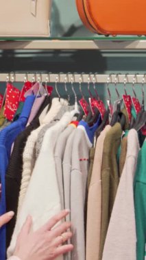 Uzun siyah saçlı şık bir genç kadın şık bir butikte şık bir elbise dizisinin ortasında yumuşak, renkli bir kazağı inceliyor. Göz alıcı renkler ve modaya uygun aksesuarlarla çevrelenmiş bir şekilde alışverişin heyecanını yayıyor.
