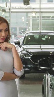 Göz kamaştırıcı genç bir kadın, pırıl pırıl yeni bir arabayı değerlendirirken kaşları dalgın dalgın, modern bir galeride duruyor. Etrafı lüks araçlar ve canlı yeşilliklerle çevrili olan kadının ifadesi,