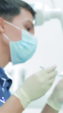 Maske ve eldiven takmış, çalışkan genç bir dişçi hastaların dişlerini titizlikle inceliyor. Parlak, steril çevre odaklı bir lamba ile geliştirilir, profesyonellik ve bakım atmosferi yaratarak,