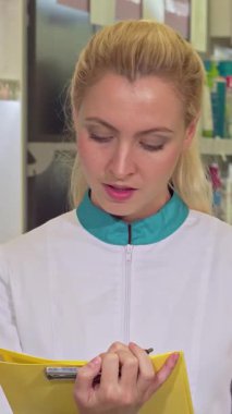 Kendini işine adamış genç bir kadın eczacı, mavi yakalı gevrek beyaz bir laboratuvar önlüğü içinde envanteri dikkatle incelerken etrafındaki renkli raflar ilaçlarla dolup taşıyor. Odaklanmış bakışları ve parlak sarı saçları bağlılığını simgeliyor.