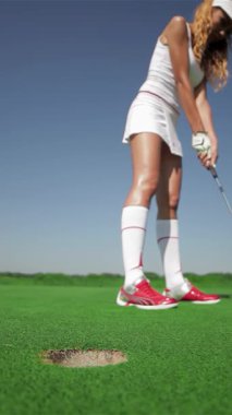 Odaklanmış genç beyaz bir kadın golf topunu delikten çıkarmak için zarifçe eğilir ve yemyeşil sahada yeteneğini sergiler. Şık beyaz bir elbiseyle giyinmiş, parlak mavi gökyüzünün altında parlak kırmızı saçları parlıyor,