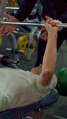 Odaklanmış Kafkasyalı üst düzey bir adam tutkulu bir şekilde göğüs egzersizi yapar, destekleyici genç bir erkek antrenör rehberlik ederken bir halteri kaldırır. Dinamik spor salonu atmosferi canlı ağırlık ve son teknoloji ekipmanlarla dolu.