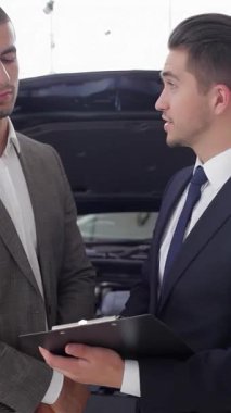 Gösterişli bir otomobil galerisinde, özel dikim takım elbiseli karizmatik genç bir satıcı çarpıcı lüks bir araçla ilgili potansiyel bir alıcıyı tutkuyla meşgul ediyor. Parlak, modern ortam heyecan ve sofistike bir ortam yaratıyor.