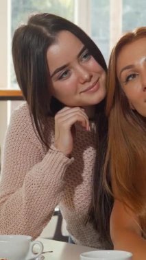 Canlı bir kahve dükkanında, birinin dalgalı kızıl saçlı, diğerinin parlak siyah bukleli iki neşeli genç kadın şakacı bir şekilde selfie çekiyorlar. Zarif hamur işleriyle bezenmiş ve yumuşak, sıcak ışıkla yıkanmış davetkar atmosfer mükemmel.