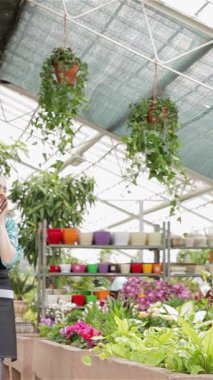 Dalgalı sarı saçlı neşeli bir çiçekçi elinde akıllı telefonuyla renkli çiçekliklerden oluşan bir çiçek tarhında zarif bir şekilde geziniyor ve canlı bir sohbete dalıyor. Şık bir önlük giymişti. Sevgiyle yemyeşil bitkileri spreyliyordu.